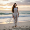 Woman in a white macramé dress walking on a beach at sunset.