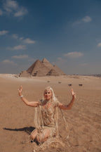   Woman seated on desert sand in a gold silk goddess gown, Pyramids of Giza behind her — Hathor