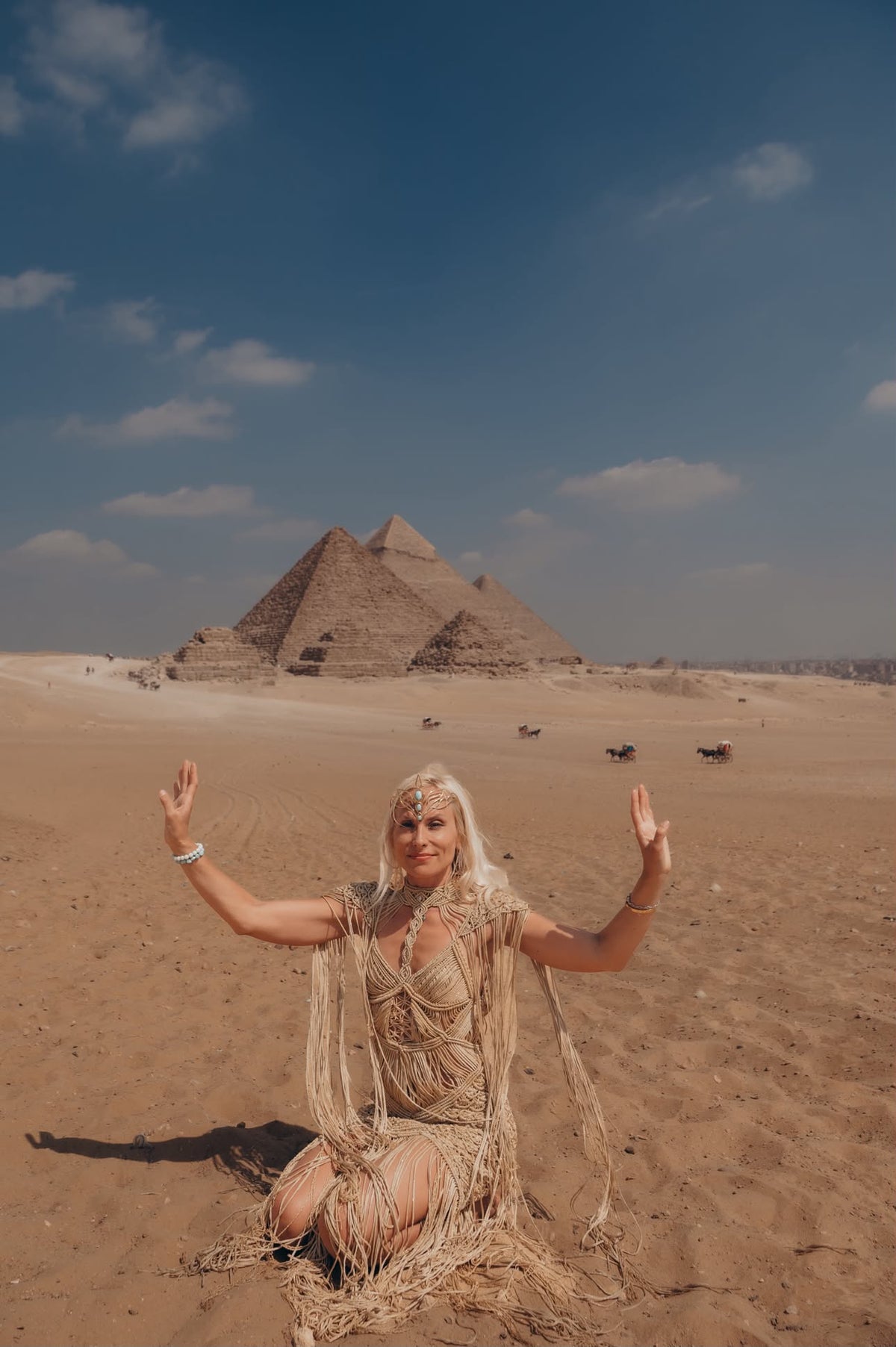   Woman seated on desert sand in a gold silk goddess gown, Pyramids of Giza behind her — Hathor