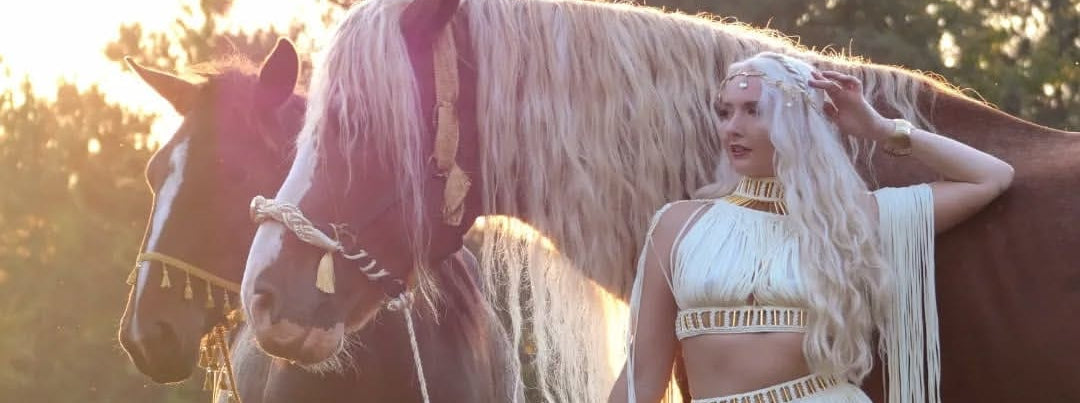 Woman in a white fringe outfit standing next to a horse in a field with trees in the background.