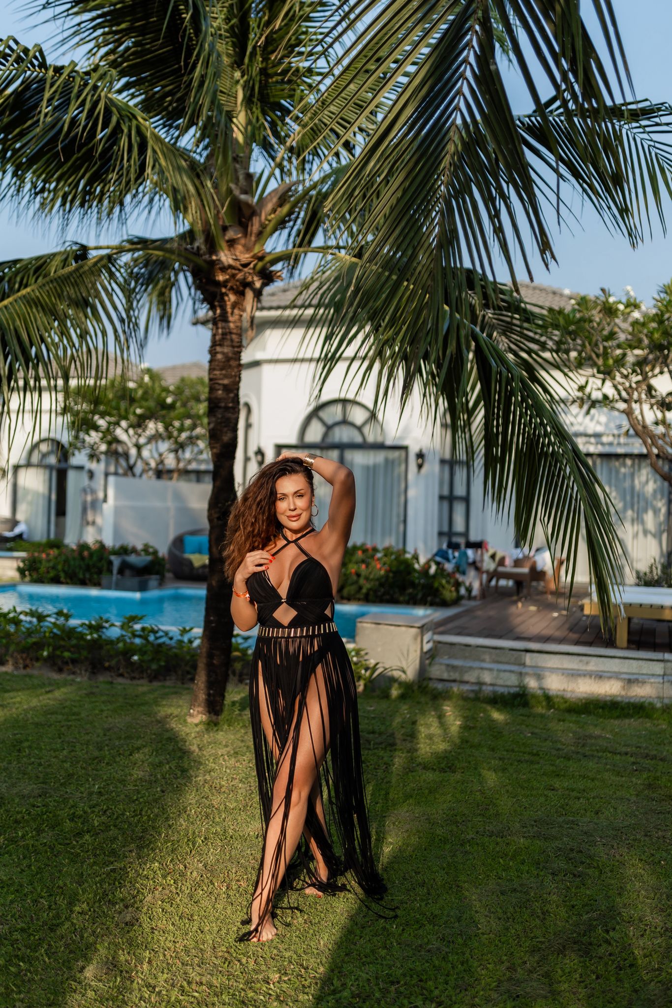A woman in a black fringe dress stands in a tropical setting with a palm tree and a swimming pool in the background. The dress has a halter neckline and a high slit, with a decorative belt. The background includes a white building.